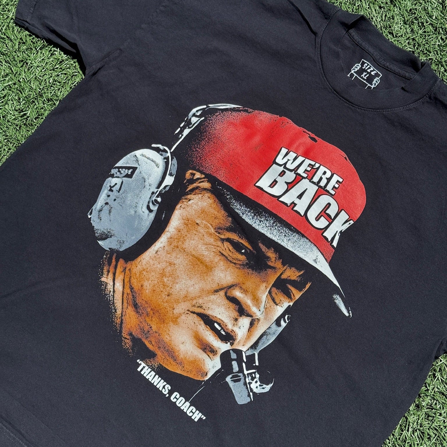 Black t-shirt with a graphic of a person wearing a red cap with 'We're Back' text, on grass.