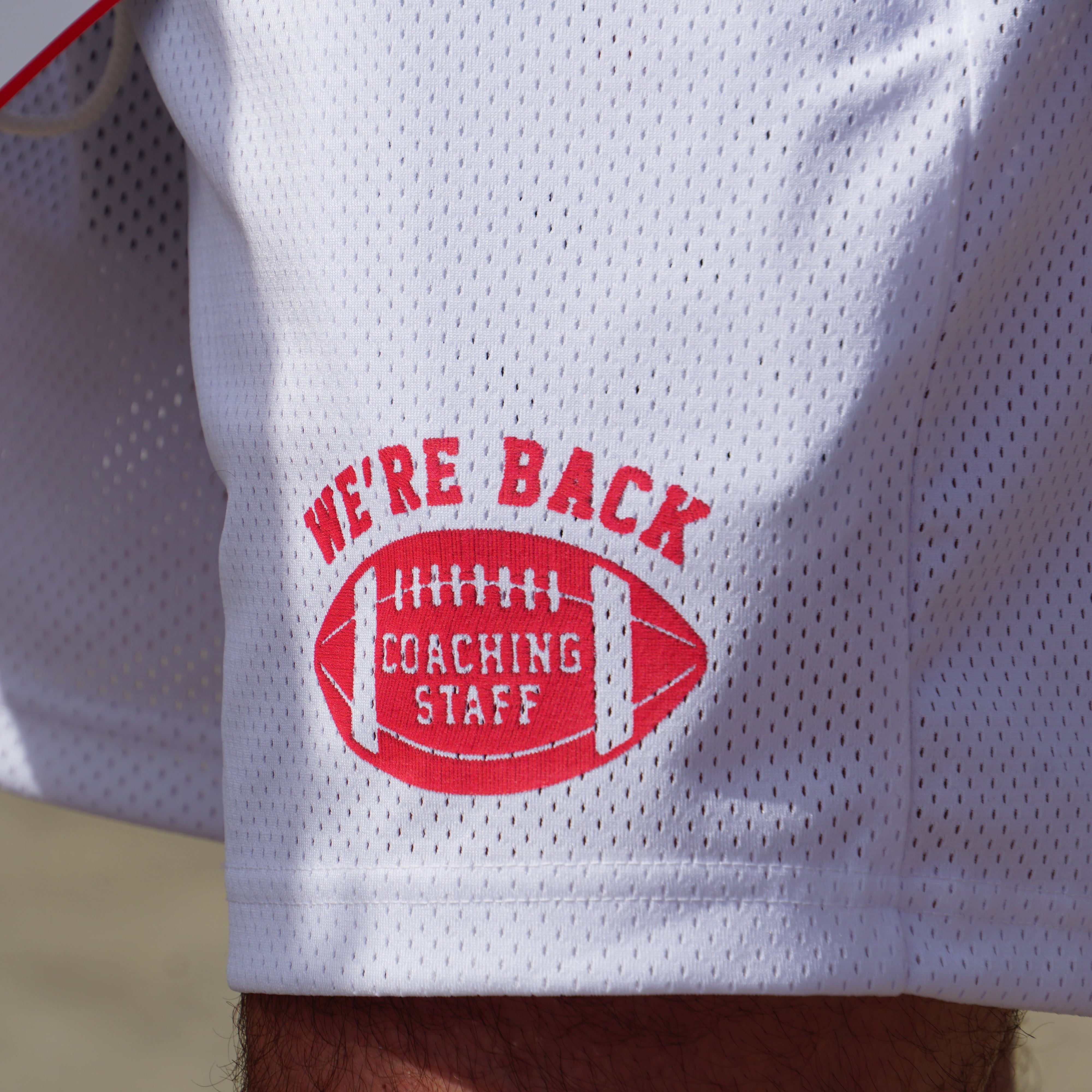 White sports jersey with red 'We're Back' and 'Coaching Staff' logo on a blurred background