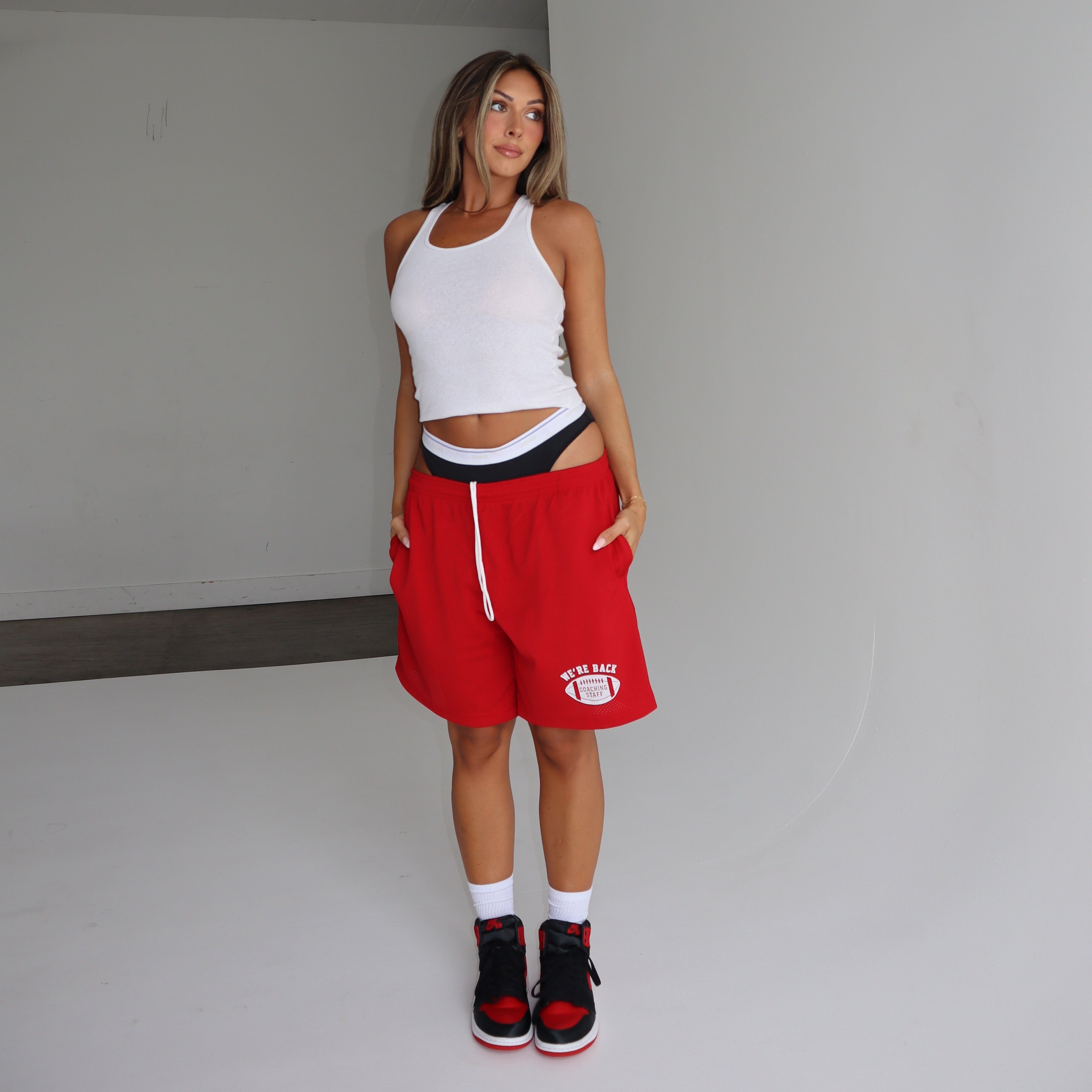 Woman wearing a white tank top and red shorts standing in a minimalistic room.