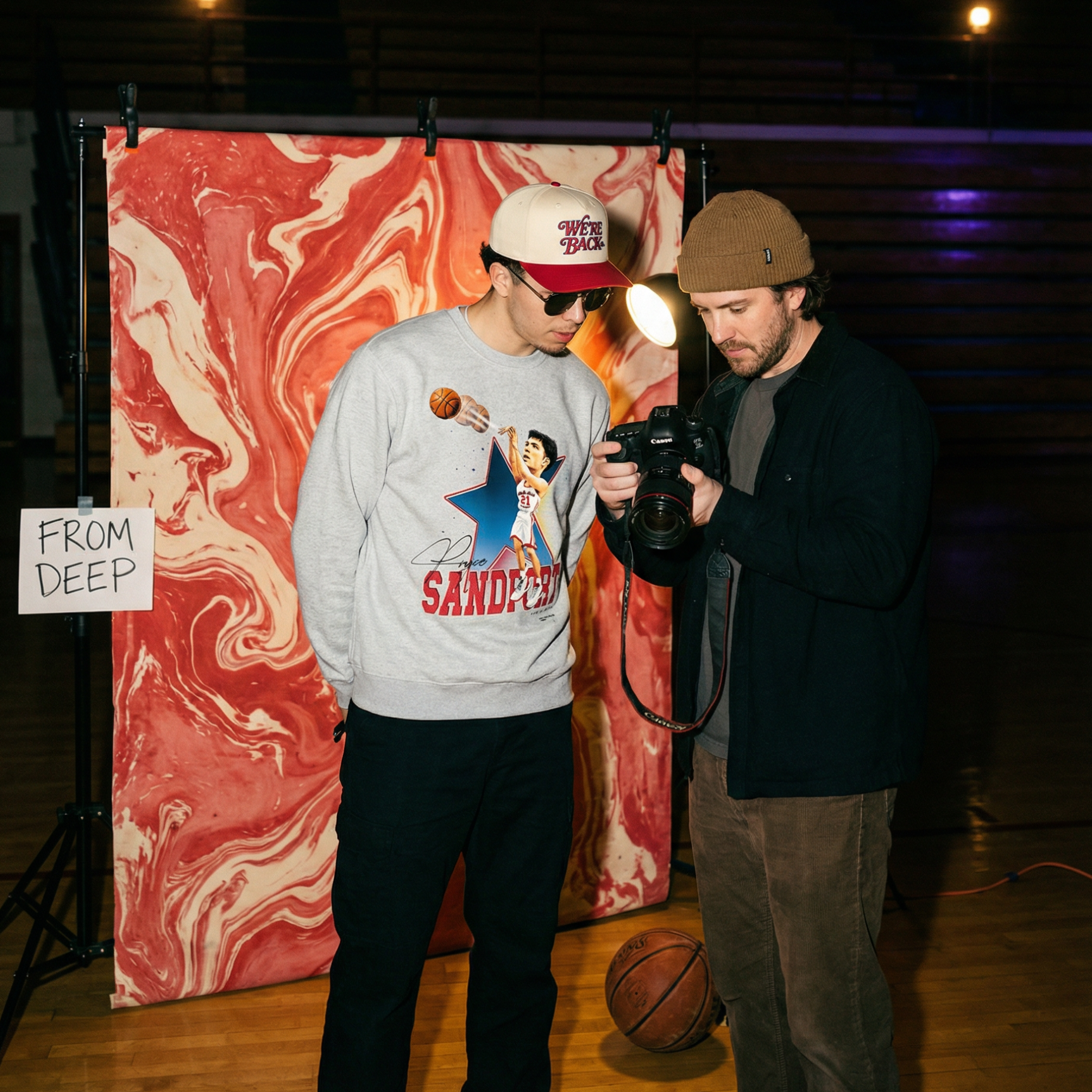 Two men posing with a basketball and camera in front of a marbled backdrop