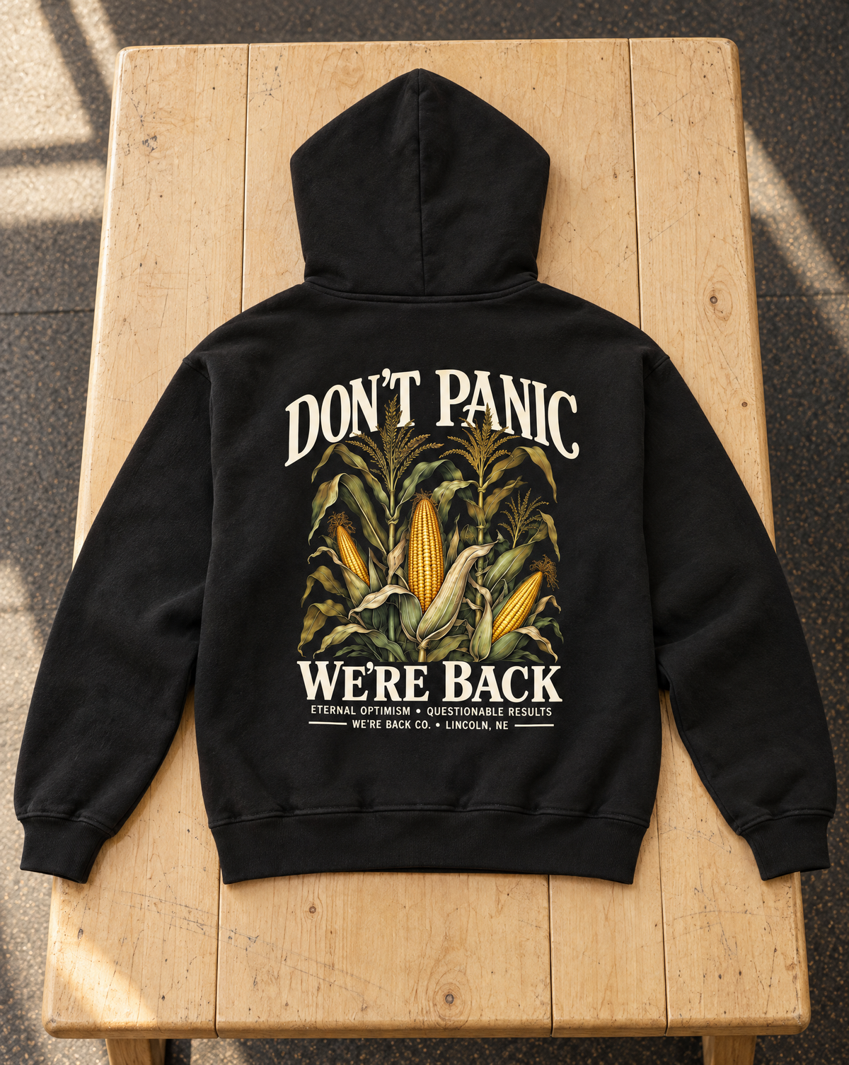 True Black Don’t Panic Hoodie laid flat on a pale wood bench, showing cream back lettering and cornfield artwork