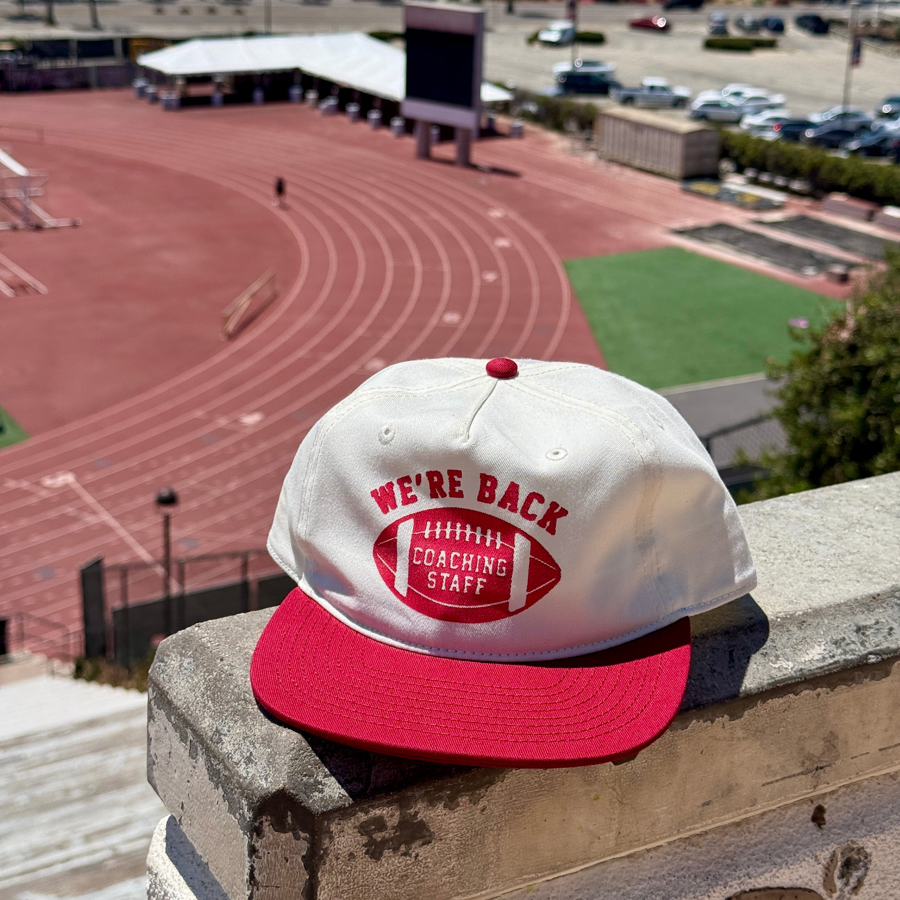 White cap with red brim and text on a ledge overlooking a track and field with palm trees and ocean in the background.