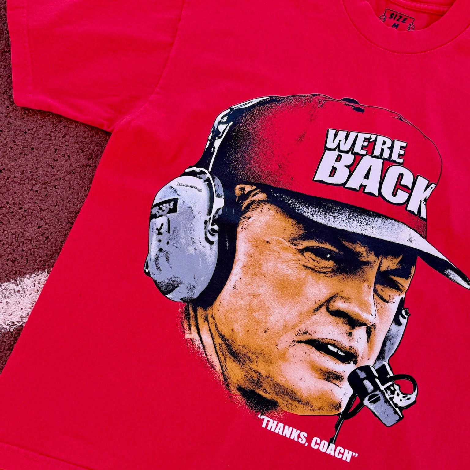 Red t-shirt with a graphic of a person wearing a cap and headphones, with text 'WE'RE BACK' and 'THANKS, COACH'.