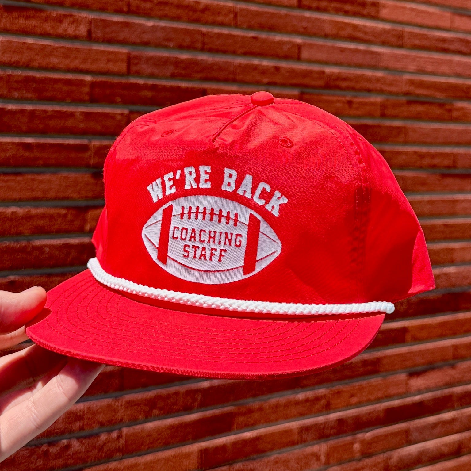 Red cap with 'We're Back Coaching Staff' text held against a brick wall.