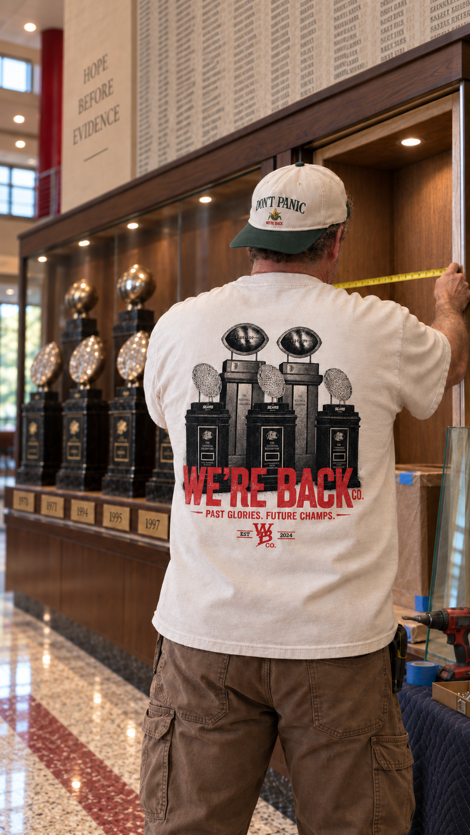 Fan wearing the Trophy Tee measures an empty space beside five championship trophies.